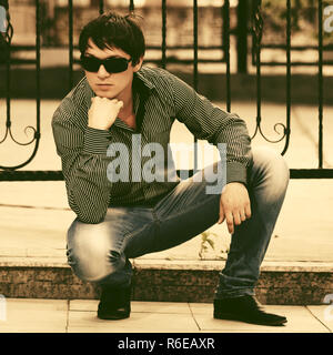 Young handsome man in sunglasses and striped shirt sitting on sidewalk in city street Stock Photo