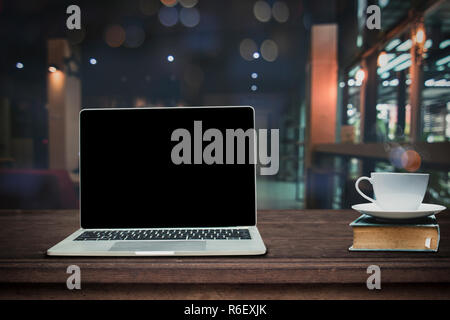 Hot cup of coffee on big book and laptop on wood table in front of blurred cafe restaurant background Stock Photo