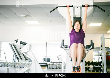Young brunette woman is engaged in fitness in the gym. Performs physical exercises for the muscles of the press, bending his legs to his chest, hanging on a horizontal bar Stock Photo