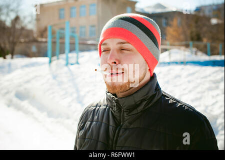 Portrait of a man with a beard with a mustache, smoking a cigarette, with an emotional face in the open air. Dressed in a bright striped hat, against the background of a snowy sunny day Stock Photo