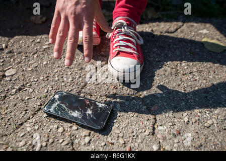 Broken and damaged smartphone with cracks on glass, screen laying on ground. Young man is disappointed and sad, picking up phone and holding in hands. Stock Photo