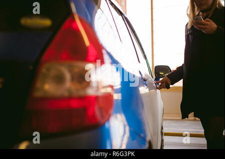 Woman in parking lot on blue car background. Opens door with key Stock Photo