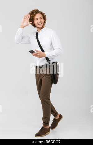 Full length portrait of a cheerful businessman dressed in shirt isolated over white background, talking on mobile phone, waving hand Stock Photo