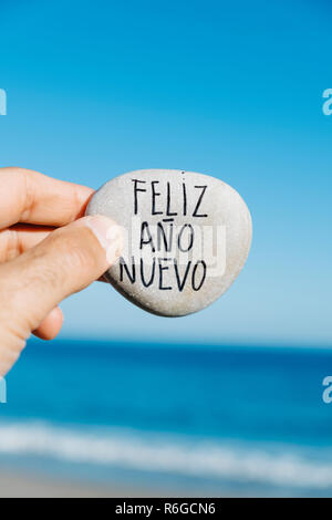 closeup of a young man on the beach, with a stone in his hand, with the text feliz ano nuevo, happy new year written in spanish, and the ocean in the  Stock Photo
