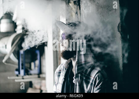 young man with beard in light checkered jacket with hood in a cap smokes an electronic cigarette in vape bar Stock Photo