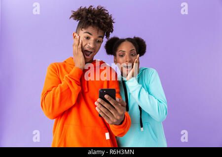 Photo of surprised african american brother and sister wearing colorful sweatshirts using mobile phone isolated over violet background Stock Photo