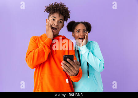 Photo of shocked african american brother and sister wearing colorful sweatshirts using mobile phone isolated over violet background Stock Photo