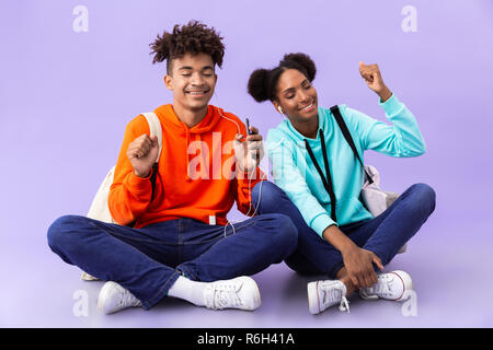 African american couple wearing backpacks using mobile phone and earphones while sitting on floor with legs crossed isolated over violet background Stock Photo