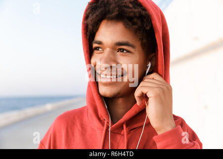 Close up of a smiling young african teenager dressed in hoodie standing at the beach, listening to music with earphones Stock Photo