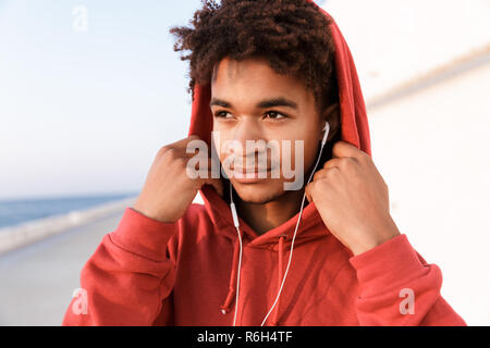 Close up of a young african teenager dressed in hoodie standing at the beach, listening to music with earphones Stock Photo