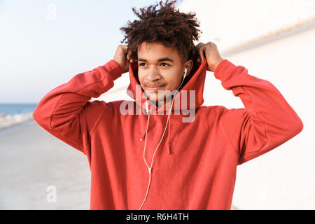 Close up of a handsome young african teenager dressed in hoodie standing at the beach, listening to music with earphones Stock Photo