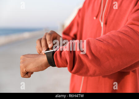 Close up of a young african teenager dressed in hoodie standing at the beach, using smart watch Stock Photo