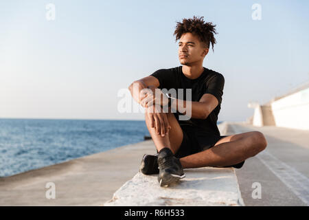 Image of concentrated young guy sportsman sitting outdoors on the beach. Stock Photo