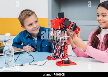 adorable schoolgirl and smiling schoolboy sitting at desk and working together on robot model at STEM class Stock Photo