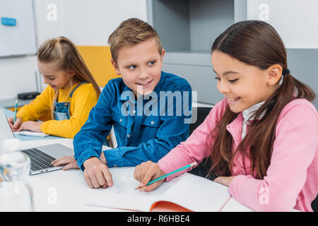 schoolboy and schoolgirls writing in notebooks and using laptop during stem lesson Stock Photo
