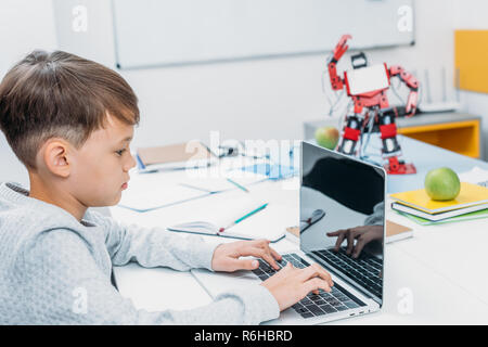 concentrated schoolboy typing on laptop keyboard and programming mechanical robot in stem class Stock Photo