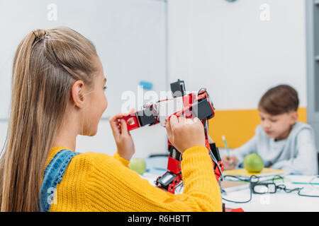 schoolgirl sitting at table and holding robot model during STEM lesson with classmate on background Stock Photo