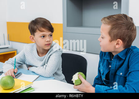 schoolboys sitting at desk, having break, talking and eating apples in classroom Stock Photo