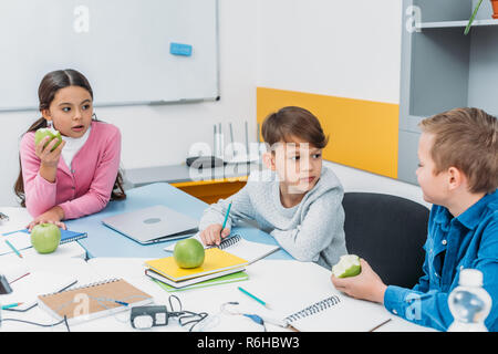 classmates sitting at table, having break, talking and eating apples in classroom Stock Photo