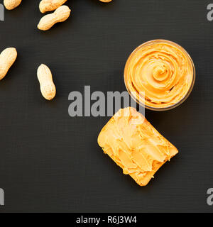 Bowl of peanut butter, toast, unshelled peanuts over black surface, overhead view. From above. Stock Photo