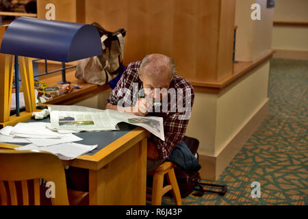 Older man reading, studying newspaper with magnifying glass at desk in public library Stock Photo