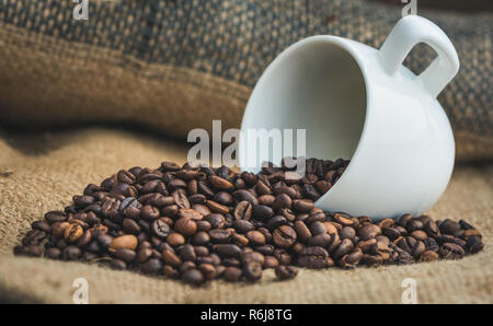 Roasted Coffee Beans Spilled out of White Ceramic Cup on Burlap Background Stock Photo