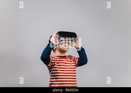 Excited kid having fun with VR glasses. Portrait of cheerful child wearing virtual reality headset against grey background. Stock Photo