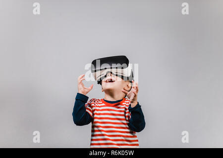 Excited kid having fun with VR glasses. Portrait of cheerful child wearing virtual reality headset against grey background. Stock Photo