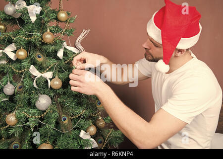 Man in santa hat decorate Christmas tree with balls, bows, garland. xmas, new year, eve, holidays preparation, celebration. Festive decorations and ornaments concept Stock Photo