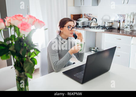 Woman talking on phone and drinking tea while working on laptop on modern kitchen. Freelancer relaxing Stock Photo