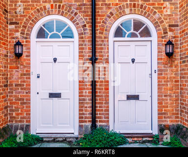 Two white wood residential front doors, with a drain pipe down the middle. The walls are red brick there are lunette arches over the doors and metal l Stock Photo