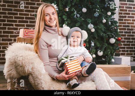 Theme Christmas holidays winter new year. A young stylish Caucasian mother holds her son in her arms for 1 year in a funny shirt sitting on a sleigh and wearing fur under the tree of decorated house Stock Photo