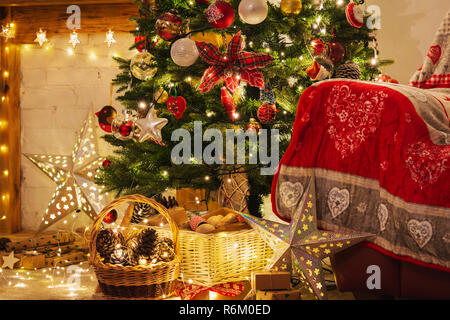 Christmas living room, decorated fireplace with wood mantelpiece, lit up Christmas tree with baubles, stars, pine cones, cosy armchair with read throw Stock Photo