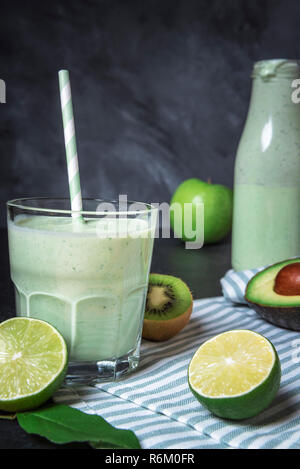 Green smoothie in a glass, surrounded by limes, kiwi, avocado, and apples, on a kitchen table with a towel. Healthy lifestyle concept. Detox drink. Stock Photo
