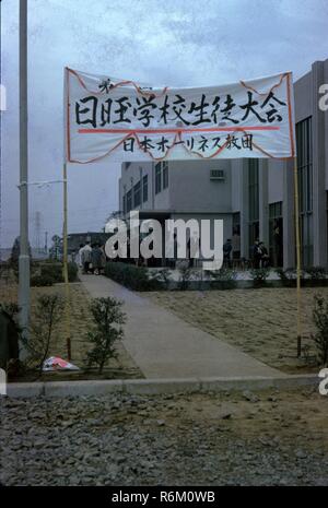 Color photograph of a group of Japanese people standing outside, near the entrance to a concrete, Art Deco style building (possibly a church or hospital) with a sign written in Japanese characters attached to bamboo poles, and a sign with a red cross visible in the left foreground, likely photographed in Japan during the mid-twentieth century, 1965. () Stock Photo