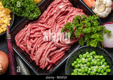 Raw minced meat in container with ingredients for shepherds pie with green peas, yellow corn, carrot, onion and seasonings, on black background, top v Stock Photo