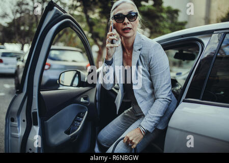 Senior businesswoman getting out of a car talking on phone. Female entrepreneur travelling to office in a luxurious car. Stock Photo