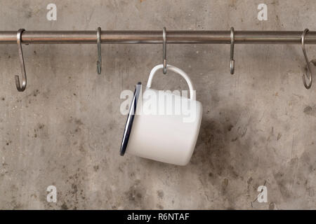 White tin cup hanging on stainless rail on cement wall background. Stock Photo