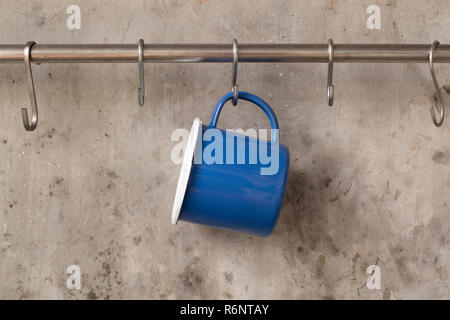 Blue tin cup hanging on stainless rail on cement wall background. Stock Photo