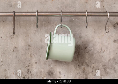 Green tin cup hanging on stainless rail on cement wall background. Stock Photo