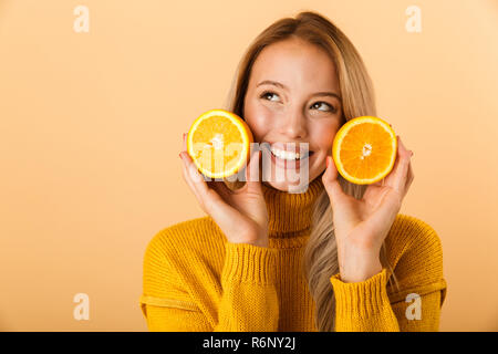 Portrait of a smiling young woman dressed in sweater standing isolated over yellow background, holding sliced orange at her face Stock Photo
