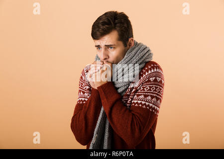 Portrait a young man dressed in sweater and scarf isolated over beige background, warming up arms Stock Photo