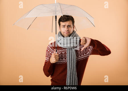Portrait a sad young man dressed in sweater and scarf isolated over beige background, standing under umbrella, pointing thumb down