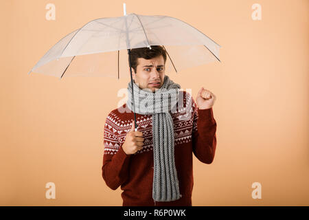 Portrait a sad young man dressed in sweater and scarf isolated over beige background, standing under umbrella