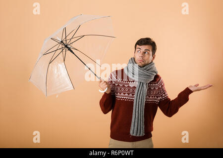 Portrait a confused young man dressed in sweater and scarf isolated over beige background, standing under umbrella