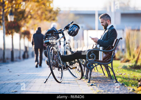 A businessman commuter with bicycle sitting on bench in city and using smartphone when traveling home from work. Stock Photo