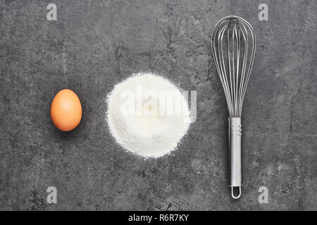 Egg, flour and whisk on dark grey concrete background. Ingredients for dough or noodles preparation. Top view. Copy space. Stock Photo