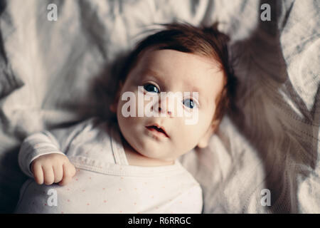 Closeup portrait of cute adorable funny white Caucasian brunette little baby newborn with blue grey eyes lying on bed looking in camera. Authentic lif Stock Photo