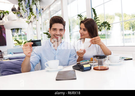 Image of a young happy loving couple sitting in cafe holding credit card and check. Stock Photo