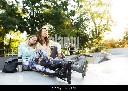 Image of happy two girls friends sisters on rollers sitting in park outdoors listening music by earphones. Stock Photo
